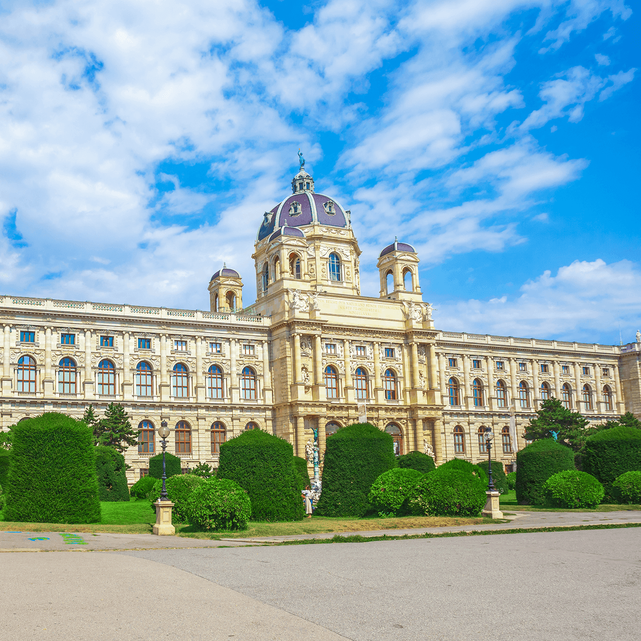 Ornate historic building with a large dome, intricate facade, and green landscape under blue sky.