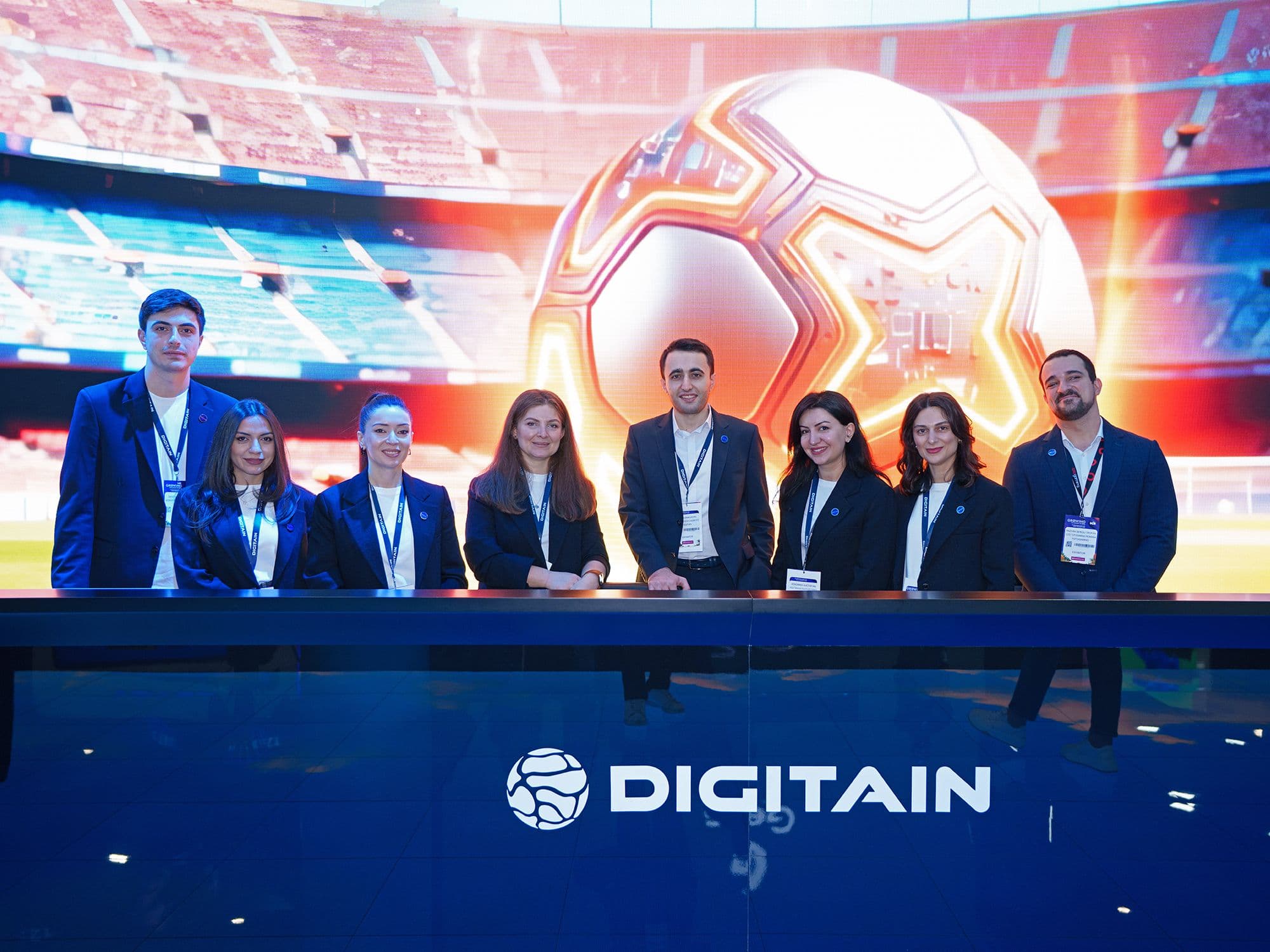Eight people behind a table with a Digitain logo, soccer stadium screen in background.