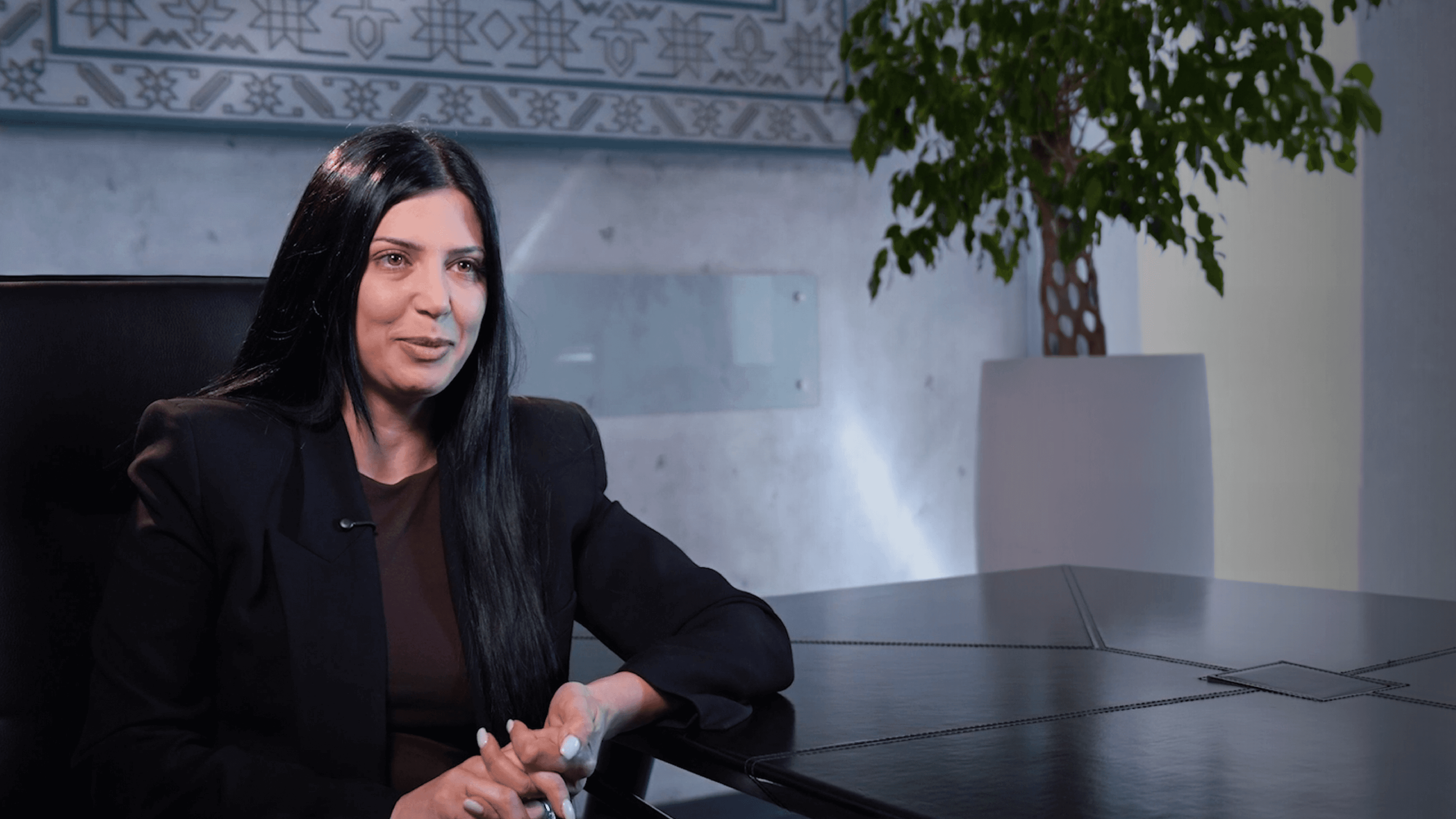 Professional woman with long dark hair sits at an office table.