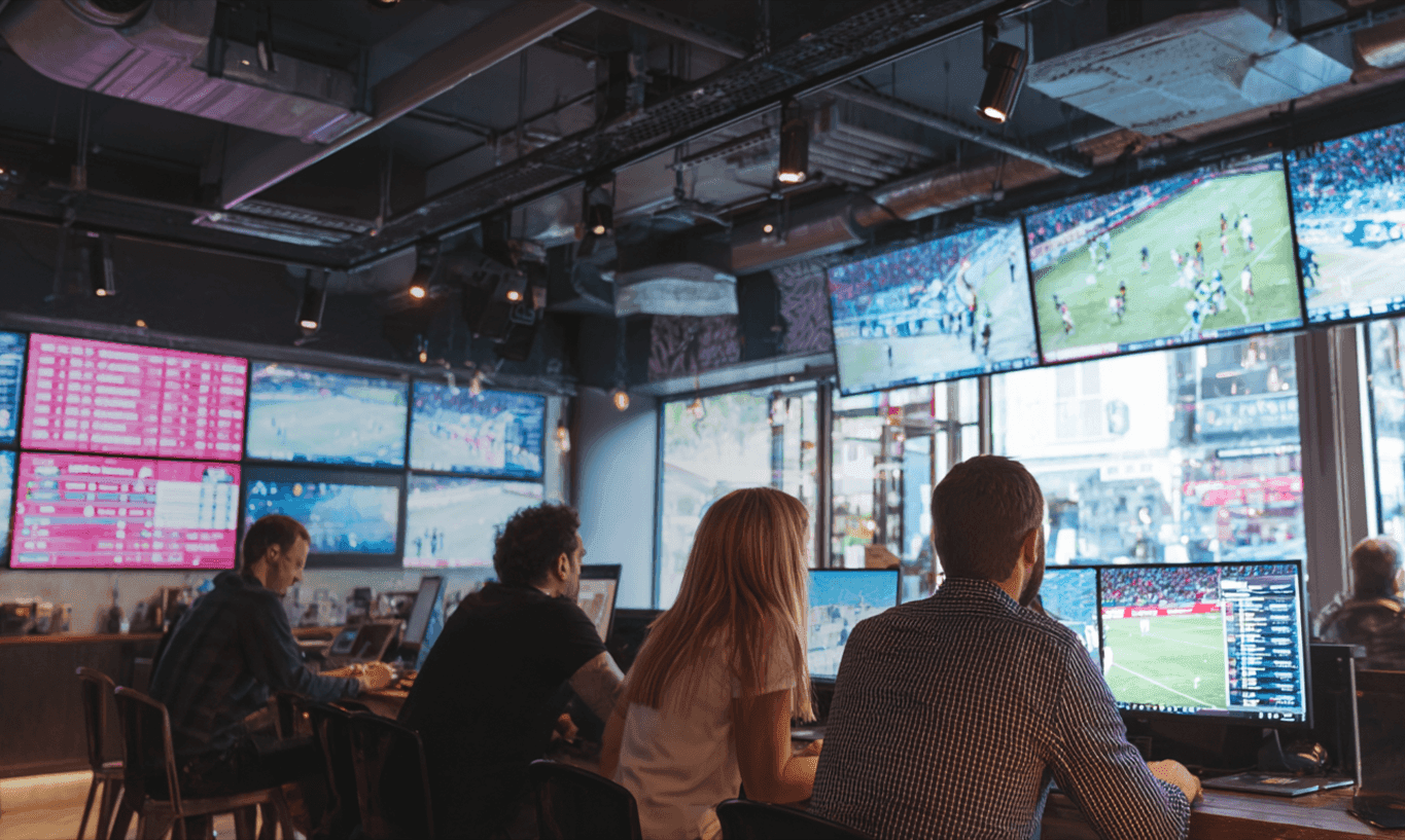 People watching multiple large screens displaying sports and data in a dimly lit room.
